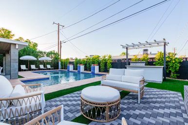 Modern residential backyard with rectangular swimming pool, poolside loungers and umbrellas, pergola-covered outdoor bar and white wicker patio seating on gray pavers