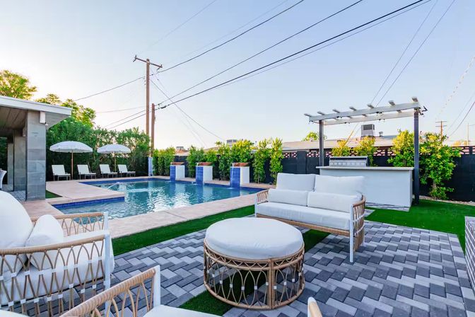 Modern residential backyard with rectangular swimming pool, poolside loungers and umbrellas, pergola-covered outdoor bar and white wicker patio seating on gray pavers
