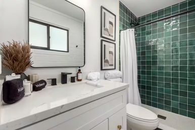 Modern bathroom with white vanity and marble countertop, matte black faucet and framed mirror, rolled white towels and dried pampas decor, and a deep green tiled shower with a white curtain.