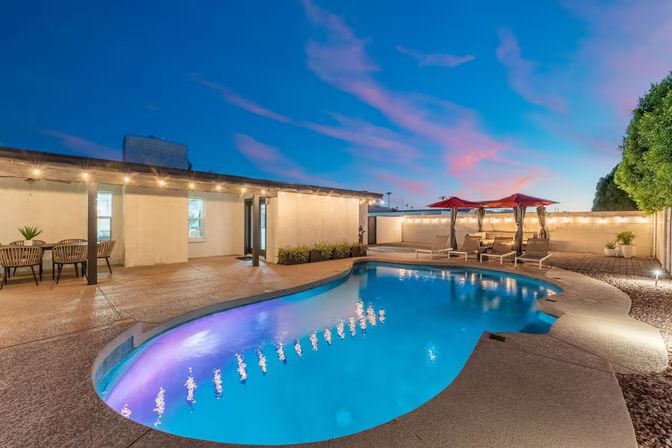 Twilight backyard oasis with a blue-lit freeform swimming pool, concrete deck and covered patio strung with lights, outdoor dining area and loungers with red umbrellas.