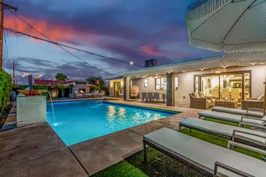 Inviting suburban backyard at dusk with an illuminated rectangular swimming pool, cushioned lounge chairs, covered patio dining and wicker seating, outdoor fireplace and colorful sunset sky.