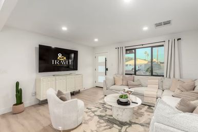 Bright modern living room with neutral sectional sofa, plush accent chair, round marble coffee table, wall-mounted flat-screen TV, potted cactus, large window overlooking the neighborhood, light hardwood floors and patterned area rug.