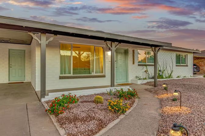 Sunset-lit single-story ranch house with covered porch, mint-green front door and warm-lit window, curved path lined with solar lights through a low-maintenance gravel desert garden with flowering shrubs, agave and barrel cactus.