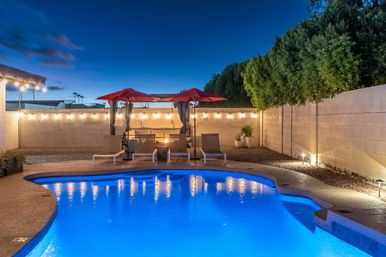 Dusk backyard pool with vibrant blue-lit water, lounge chairs and red umbrellas around a warm fire pit, string lights along a privacy wall and manicured trees.