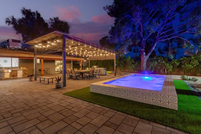 Modern backyard patio at dusk with a pergola draped in string lights, outdoor dining area and kitchen, and a tiled illuminated plunge pool beside a large tree