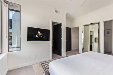 Sunlit modern bedroom with white walls, wall-mounted TV, tall window, mirrored closet doors, white bed and patterned rug