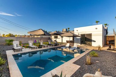 Sunlit backyard swimming pool with in-pool loungers, several chaise lounges and scalloped umbrellas on a travertine patio beside a modern white one-story home, gravel landscaping and palm trees under a clear blue sky.