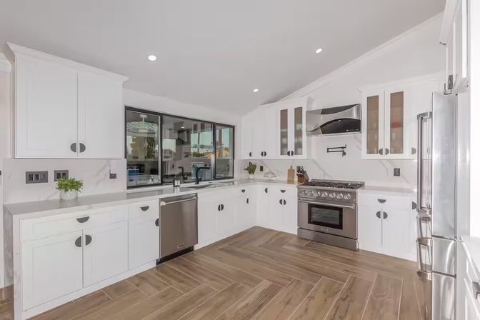 Bright, open modern white kitchen with stainless steel appliances, gas range, dishwasher, shaker cabinets, large window and wood-look tile floor