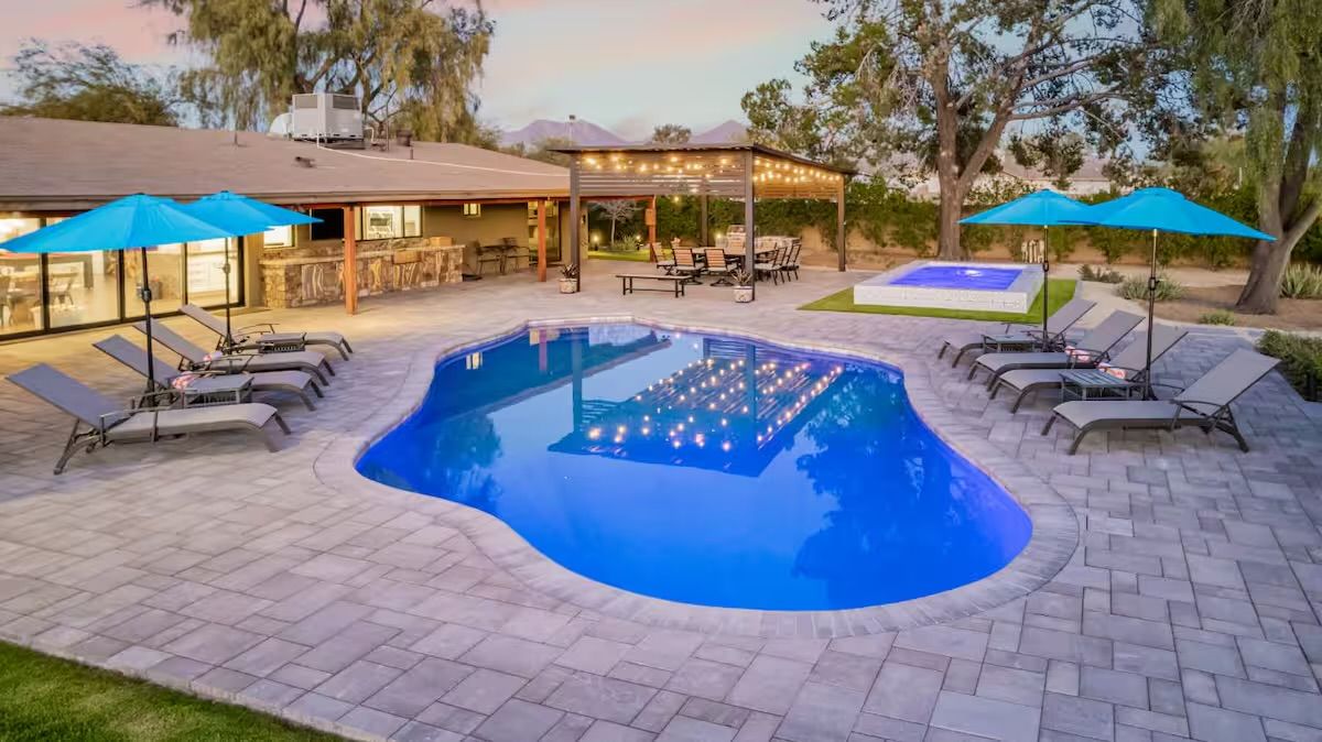 Curved blue backyard pool at dusk on a paver patio with lounge chairs, turquoise umbrellas, a string-lit pergola and raised spa — outdoor entertaining space.