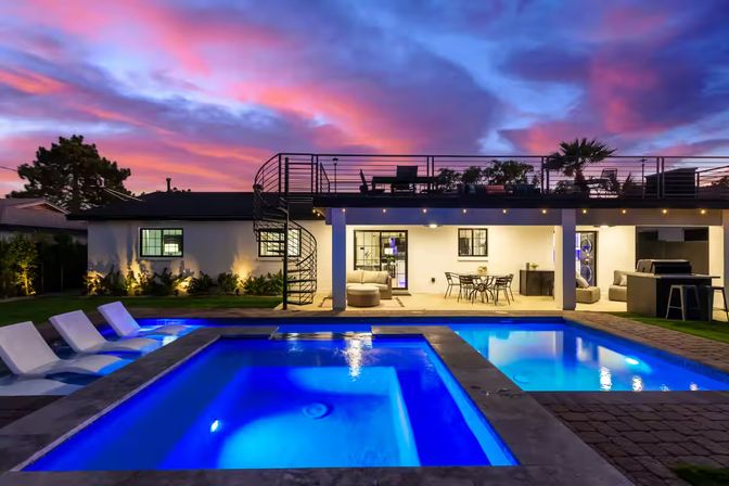 Modern backyard at sunset with glowing blue pool and spa, lounge chairs, spiral staircase to a rooftop deck, outdoor dining and grill.