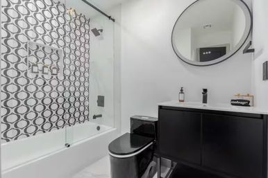 Modern black-and-white bathroom featuring a bathtub with glass sliding door, bold geometric circle-pattern tile, black toilet and sleek black vanity topped with a round mirror and white countertop.