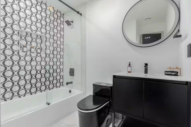 Modern black-and-white bathroom featuring a bathtub with glass sliding door, bold geometric circle-pattern tile, black toilet and sleek black vanity topped with a round mirror and white countertop.