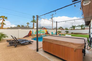 Sunny Southwestern backyard with covered hot tub, small freeform pool, black lounge chairs, string lights, palm trees, and a colorful desert mural on a white block wall.