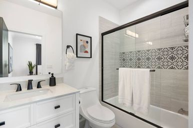 Bright modern bathroom with white vanity and sink, matte black faucets, toilet, glass sliding shower door with decorative patterned tile accent strip and white towels.