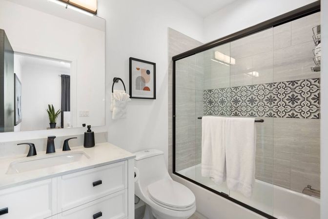 Bright modern bathroom with white vanity and sink, matte black faucets, toilet, glass sliding shower door with decorative patterned tile accent strip and white towels.