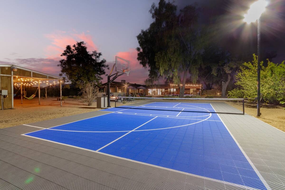 Blue outdoor multi-sport court with basketball hoop and tennis net lit by a tall floodlight at dusk, pink sunset sky, string-lit patio, hammocks and large trees near a house in the background.