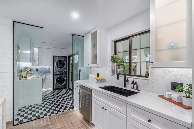 Bright modern home laundry and utility room with white cabinets, marble-look countertops, black undermount sink and faucet, stainless dishwasher, stacked front-load washer and dryer behind a glass partition, black-and-white checkered floor, and potted plants by a large window.