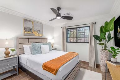 Bright modern bedroom with a wooden headboard and white linens, textured blue pillows and an orange throw, matching nightstands and lamps, abstract gold-accent artwork, ceiling fan, window with curtains, tall potted plant, and wood-look tile floor.