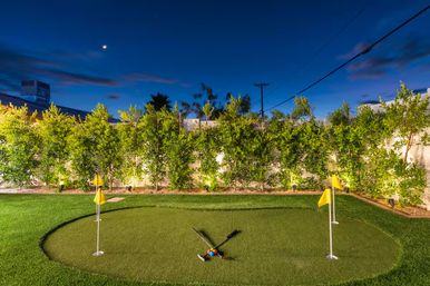 Evening backyard putting green with four yellow flags, crossed croquet mallets and colorful balls on synthetic turf, illuminated hedge lighting and a crescent moon overhead.