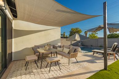 Sunny suburban backyard patio with a beige shade sail, modern wicker sofa and stools around a small dining table, lounge chairs and umbrella beside a swimming pool under a clear blue sky.