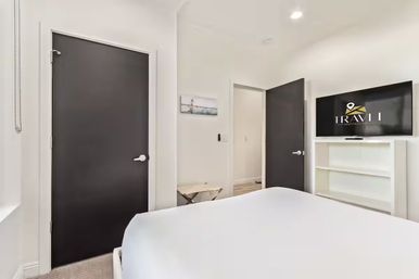 Bright modern urban apartment bedroom with white walls and linens, two dark charcoal doors, wall-mounted flat-screen TV above white shelving, minimalist decor and small bench.