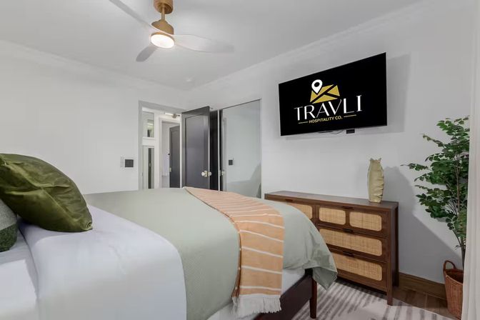 Bright modern vacation rental bedroom with king bed, green pillows and striped throw, wall-mounted TV, woven-wood dresser, ceiling fan and potted plant