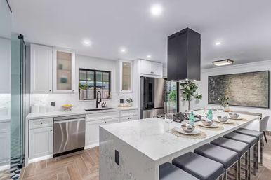 Bright modern open-concept kitchen with white cabinetry, marble island breakfast bar, stainless steel appliances, black range hood, and counter seating