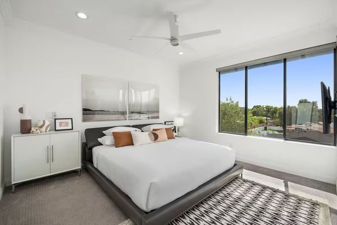 Bright modern bedroom with low black platform king bed, crisp white linens and tan accent pillows, large windows offering a sunny suburban tree-and-rooftop view, ceiling fan, abstract wall art, white storage cabinet and wall-mounted TV
