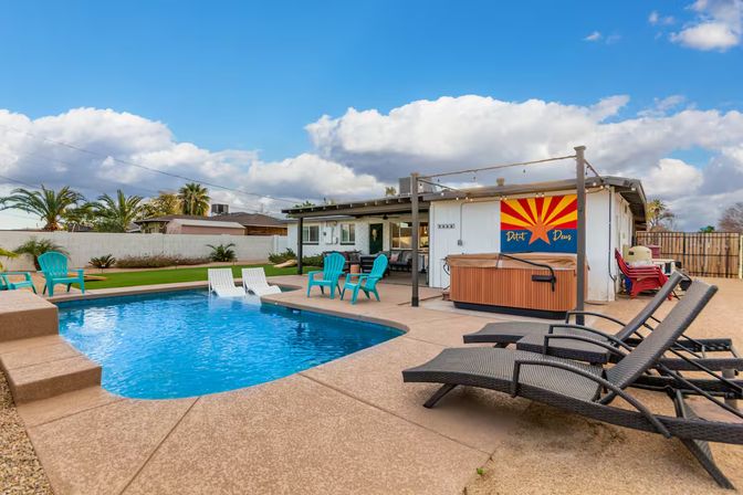 Arizona backyard pool with rectangular blue inground pool, hot tub beside a colorful sunburst mural, turquoise Adirondack chairs, in-pool loungers and black wicker chaise lounges on a concrete patio under a bright sky.