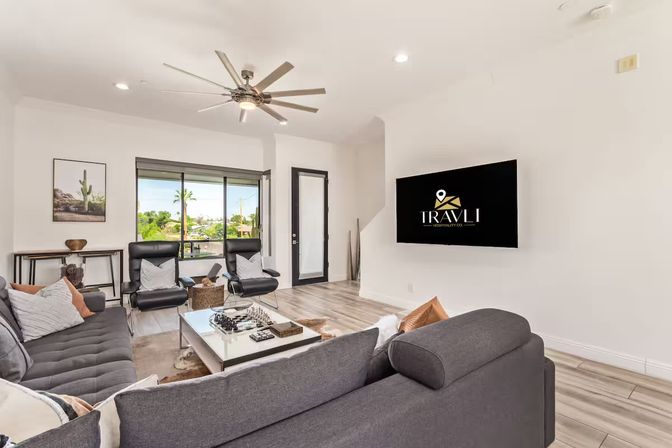 Sunlit modern living room with a gray sectional sofa, two black lounge chairs, white coffee table, wall-mounted TV, large ceiling fan, sliding glass door with palm-tree view and light wood floors.