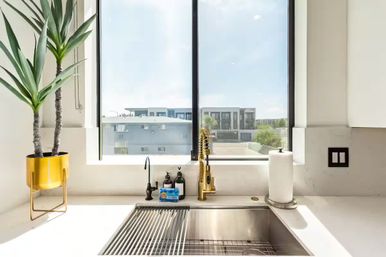 Sunlit modern kitchen sink with stainless steel basin, brass spring faucet, yellow potted plant and paper towel on a white countertop, large window overlooking urban apartments.
