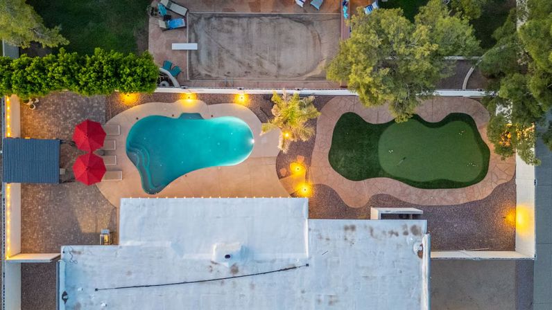 Aerial drone view of a residential backyard at dusk with a freeform turquoise swimming pool, adjacent artificial putting green, paved patio with red umbrellas, palm tree and warm string lights.