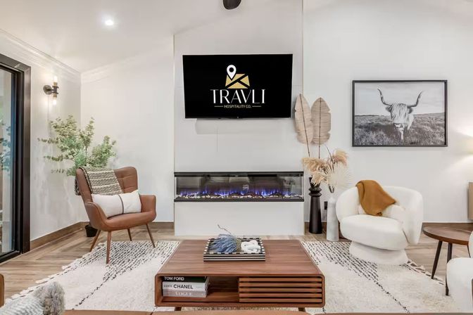 Bright contemporary living room with wall-mounted TV over a linear electric fireplace, mid-century leather accent chair, plush white swivel chair with mustard throw, wooden coffee table on a patterned rug, potted greenery and black-and-white cattle wall art — modern cozy interior.