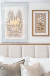 Cozy neutral bedroom interior with upholstered beige headboard, layered white and taupe pillows, and two framed wall artworks — a gold leaf abstract painting and a framed floral metal relief.