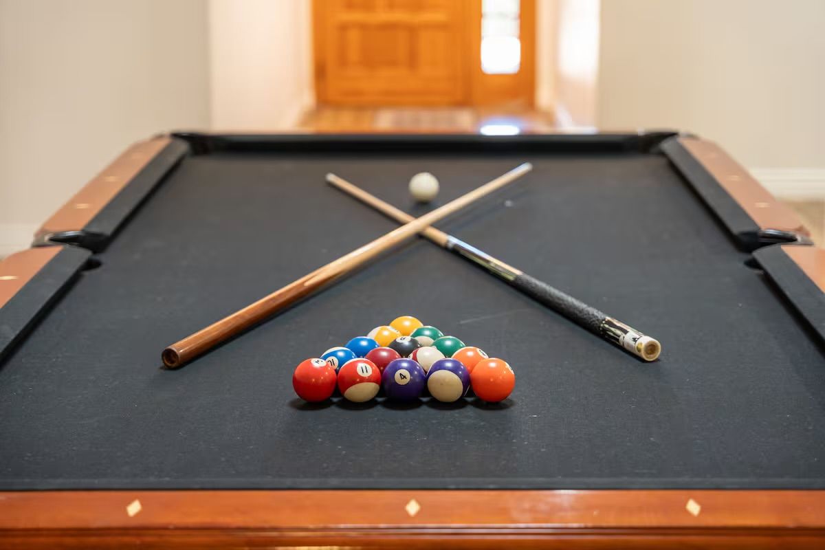 Racked colorful billiard balls on a black-felt pool table with two crossed cue sticks, white cue ball near the far rail, wooden rails and blurred doorway in an indoor game room.