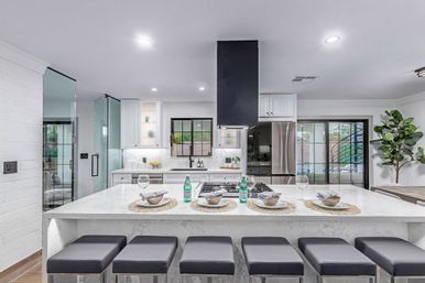 Bright contemporary kitchen with large white marble island and five gray bar stools, place settings and sparkling water, black range hood, stainless steel appliances and glass doors