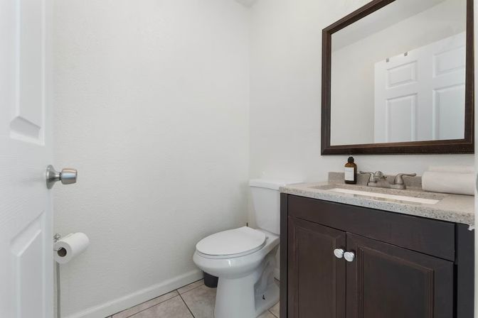 Bright, minimalist small bathroom/powder room with white toilet, dark wood vanity and quartz countertop, rectangular mirror, soap dispenser and tiled floor.