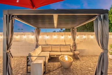Cozy backyard patio under a modern pergola — L-shaped outdoor sectional, glowing bowl fire pit, string lights and paver flooring at dusk