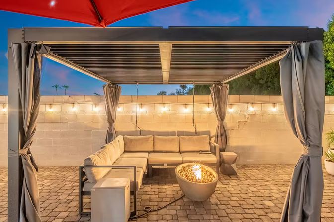 Cozy backyard patio under a modern pergola — L-shaped outdoor sectional, glowing bowl fire pit, string lights and paver flooring at dusk