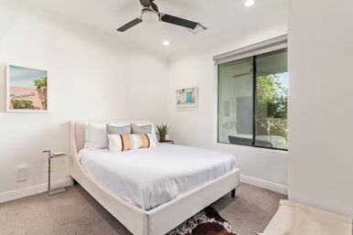 Airy modern bedroom with white upholstered bed, pastel accent pillows, ceiling fan, framed art and a large window overlooking leafy trees.