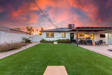 Residential backyard at a dramatic pink-orange sunset featuring a lush artificial turf lawn with cornhole boards, covered patio with outdoor seating and grill, and low landscaping.