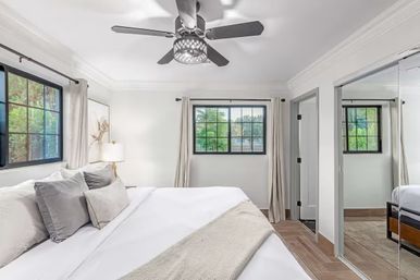 Bright modern bedroom with white linens and gray pillows, decorative ceiling fan, black‑trim windows, mirrored closet doors, and wood‑look tile floor