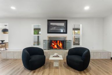 Contemporary minimalist living room with a roaring fireplace behind glass, two rounded black lounge chairs and a small marble coffee table on light hardwood floors.