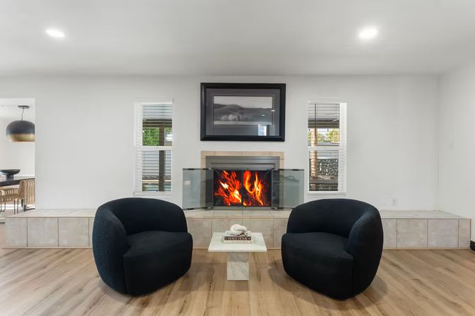 Contemporary minimalist living room with a roaring fireplace behind glass, two rounded black lounge chairs and a small marble coffee table on light hardwood floors.
