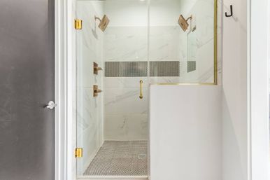 Sleek glass walk-in shower with white marble-look wall tiles, black penny-tile floor and brass fixtures