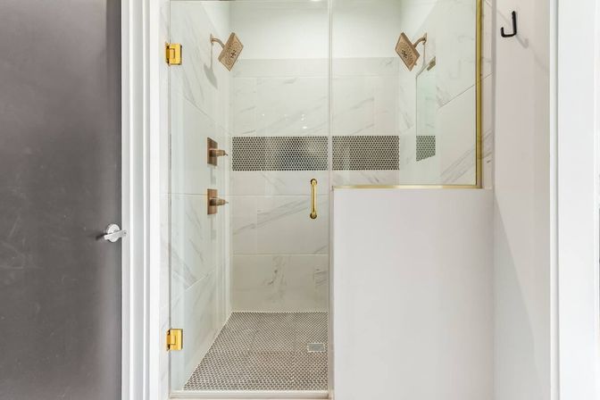 Sleek glass walk-in shower with white marble-look wall tiles, black penny-tile floor and brass fixtures