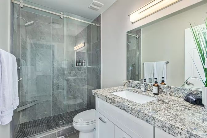 Contemporary bathroom with frameless glass walk-in shower and gray marble tile, dark mosaic shower floor, white vanity with speckled granite countertop and undermount sink, large mirror and towel accents.