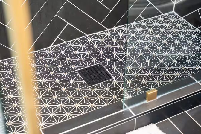 Modern bathroom shower floor with black-and-white geometric star tiles, square center drain, glass door panel and small brass accent