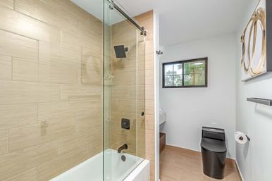 Modern bathroom with glass sliding shower door over white bathtub, beige wood-look tile, matte black shower fixtures and sleek black toilet, small window and minimalist wall art.