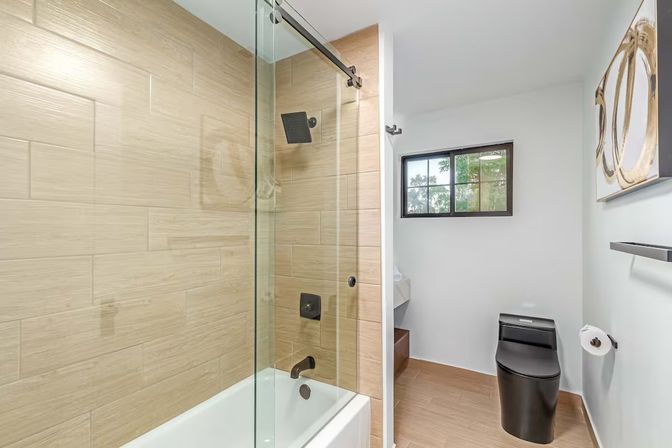 Modern bathroom with glass sliding shower door over white bathtub, beige wood-look tile, matte black shower fixtures and sleek black toilet, small window and minimalist wall art.